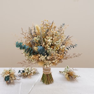 Puede incluir: Un ramo de flores secas con un peine para el cabello y un prendido a juego. El ramo presenta una variedad de flores secas en tonos beige, marrón y verde, con acentos de hojas de eucalipto verde azulado. Los tallos están envueltos con hilo.