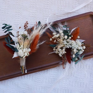 May include: Two dried flower boutonnieres with white, orange, and green accents, arranged on a wooden tray. One boutonniere is wrapped with twine and has a pearl pin. The other boutonniere is flat and has a white ribbon.
