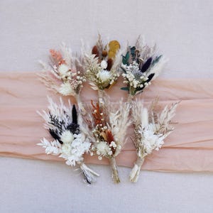 Könnte beinhalten: Sechs kleine Trockenblumensträuße in verschiedenen Farben, darunter Weiß, Creme, Orange und Schwarz. Die Stiele sind mit Bindfaden gebunden. Die Sträuße sind auf einem pfirsichfarbenen Stoff angeordnet.