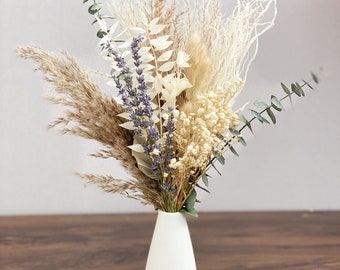 Può includere: Un vaso di ceramica bianco contiene una composizione di fiori secchi. La composizione comprende erba della pampa, lavanda ed eucalipto. I fiori sono nelle tonalità del beige, crema e viola. Il vaso ha una forma a goccia.