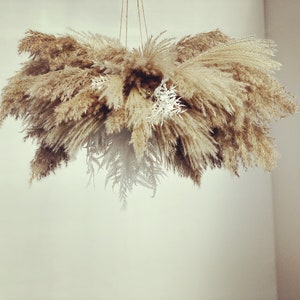 Peut inclure: Un arrangement floral séché suspendu, composé d'herbe de la pampa beige et beige clair et d'autres plantes séchées. La composition est suspendue par de la ficelle. La forme est ronde et pleine, avec un aspect doux et texturé, pour une ambiance bohème.