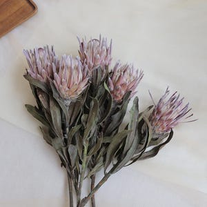 May include: A bundle of dried protea flowers with pink and lavender blooms and green leaves. The flowers have a textured appearance and are arranged on a white surface. The stems are brown and the overall aesthetic is natural and rustic.