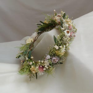 May include: A circular floral crown constructed from a variety of dried botanical elements, including small white and pink flowers, green foliage, and delicate sprigs of baby's breath, all arranged on a thin, flexible, and natural-looking vine base.