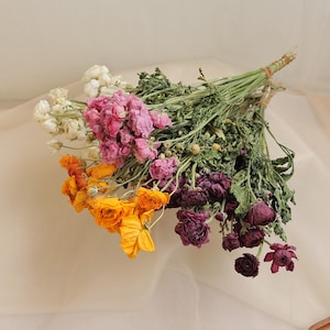 May include: A colourful bouquet of dried flowers. The arrangement includes white, pink, orange, and deep purple blooms, tied with twine. The flowers are set against a soft, neutral background.
