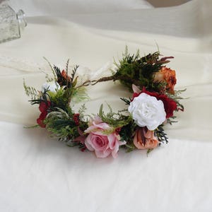 May include: A floral crown featuring a variety of flowers in shades of white, pink, red, and orange, accented with green foliage and berries. The crown is made with a woven base and a delicate ribbon.