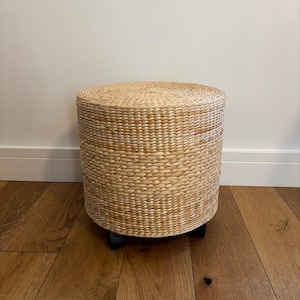 May include: A cylindrical ottoman crafted from woven light tan seagrass fibres. The piece features a flat circular top and a structured body with a consistent braided texture. It is supported by small black wheels attached to the base for mobility.