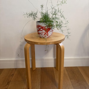May include: A round light-coloured wooden stool with four curved legs supporting a ceramic planter. The planter features a white base with a vibrant red paint-splatter pattern and contains a green asparagus fern with long, thin, trailing stems.