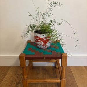 May include: A small wooden stool with four turned legs and a woven seat featuring a geometric pattern in teal and dark brown. A ceramic plant pot with a white base and red paint splatter design sits on the stool, containing a thin, wispy green plant.
