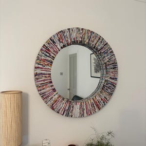 May include: A circular wall mirror featuring a wide, textured frame constructed from tightly rolled strips of colourful magazine paper. The frame displays a dense, multi-coloured pattern of various printed text and images in a radial arrangement.