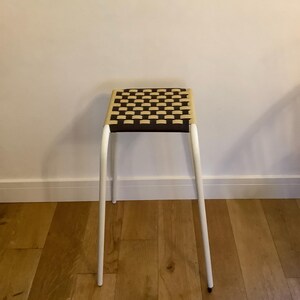 May include: A tall stool with four slender white metal legs and a square seat featuring a woven pattern of alternating dark brown and light beige fabric straps. The stool has a simple, upright frame design with a flat, grid-like woven surface.