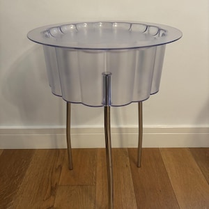 May include: A side table featuring a transparent, fluted, circular top made of clear plastic with a scalloped edge. The table is supported by three slender, metallic, silver-coloured legs that extend downward from the base of the circular top.