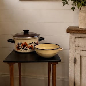 May include: A country kitchen setting with a small black table holding a floral-patterned pot and a yellow bowl with a blue rim. A sign on the wall reads "FARM FRESH GOODNESS".