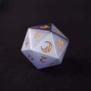 Puede incluir: Un dado poliédrico de veinte caras con un degradado de blanco a azul, con números dorados y un símbolo de luna creciente. Ideal para juegos de rol, mide aproximadamente 2,5 cm de diámetro.