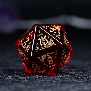 Pode incluir: Um dado vermelho de várias faces com desenhos florais e de instrumentos musicais dourados. Os números também são dourados. O dado é feito de um material translúcido e o fundo é escuro.
