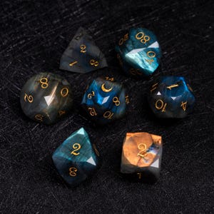 May include: A collection of seven gemstone dice in various shapes, including a d4, d6, d8, d10, d12, d20, and a cube. The dice feature a mix of blue, green, and gold colors with gold numbering. The dice are set against a dark background.
