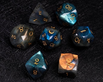 May include: A collection of seven gemstone dice in various shapes, including a d4, d6, d8, d10, d12, d20, and a cube. The dice feature a mix of blue, green, and gold colors with gold numbering. The dice are set against a dark background.