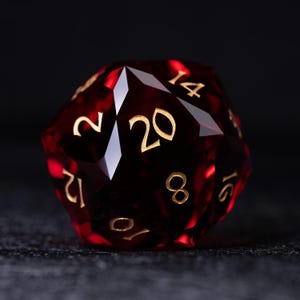 May include: A faceted, twenty-sided die in deep red with gold numerals. The die is designed for tabletop role-playing games. The number 20 is prominently displayed, with other numbers visible around the die's surface.