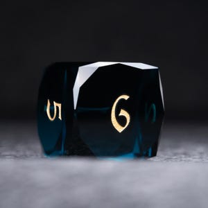 May include: A dark blue, faceted, six-sided dice with gold numbers. The dice is showing the number six.
