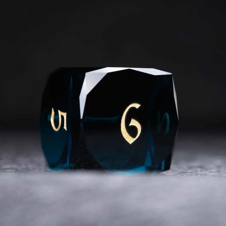 May include: A dark blue, faceted, six-sided dice with gold numbers. The dice is showing the number six.