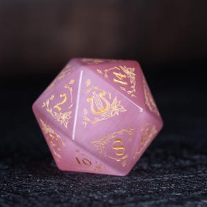 Può includere: Un dado rosa a più facce con numeri dorati e decorazioni floreali e di strumenti musicali. Il dado è realizzato in un materiale traslucido e poggia su una superficie scura e strutturata. Accessorio di gioco.