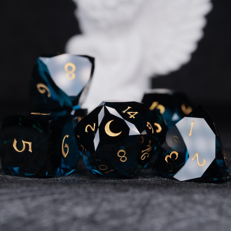 May include: A set of six blue and black polyhedral dice with gold numbers and a crescent moon symbol. The dice are made of a clear material with a dark blue tint.