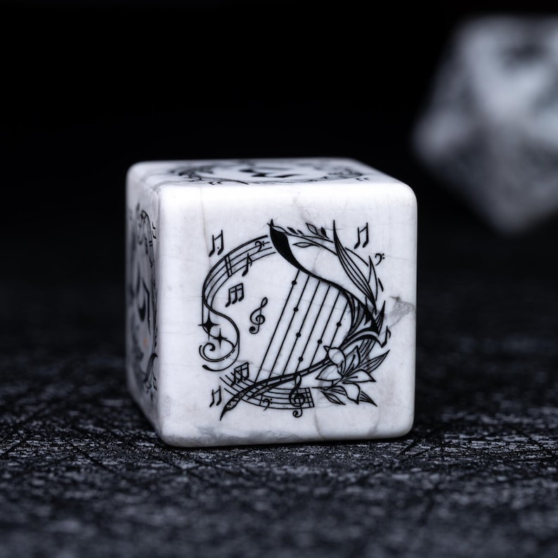 May include: A white, marble-textured die with black musical note and harp illustrations. The die features a detailed design of a harp, musical notes, and floral accents. The die is set against a dark background.