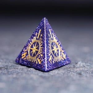 Op de afbeelding: Een paarse en gouden piramidevormige dobbelsteen met een kompasontwerp op elke zijde. De dobbelsteen is gemaakt van een glinsterend materiaal.