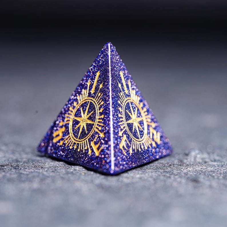 Op de afbeelding: Een paarse en gouden piramidevormige dobbelsteen met een kompasontwerp op elke zijde. De dobbelsteen is gemaakt van een glinsterend materiaal.