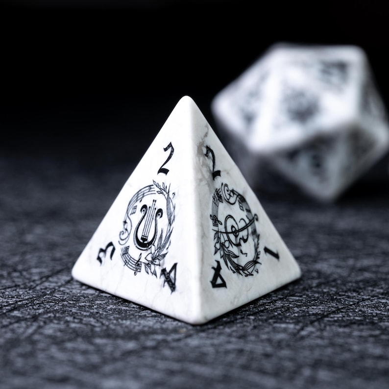 May include: A white marble-textured pyramid-shaped die with black musical note and lyre illustrations. The die has the numbers 1, 2, 3, and 4 on its faces. Another die is slightly out of focus in the background.