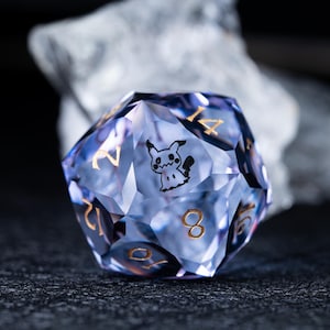 May include: A translucent, light purple twenty-sided die featuring gold-colored numbers and a black graphic of a small, ghost-like creature with pointed ears on one of its triangular faces. The die has sharp, geometric edges and a polished finish.
