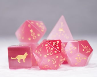 May include: A set of pink and white dice with gold cat silhouettes and paw prints. The dice are made of a translucent material and have a gradient color effect. The numbers on the dice are also in gold.
