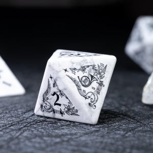 May include: A white marble-textured, six-sided die with black musical-themed illustrations. The die features a number 2, musical notes, a horn, and floral accents. The die is set against a dark, textured surface.