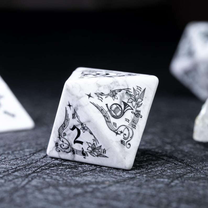 May include: A white marble-textured, six-sided die with black musical-themed illustrations. The die features a number 2, musical notes, a horn, and floral accents. The die is set against a dark, textured surface.