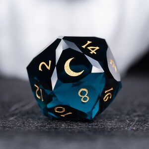 May include: A deep blue, faceted, crystal d20 dice with gold numbers and a crescent moon symbol. The dice is a 20-sided die used in tabletop role-playing games.