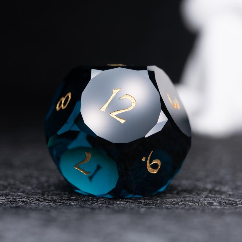 May include: A deep blue, faceted, twelve-sided die with gold numbers. The die is showing the number 12.