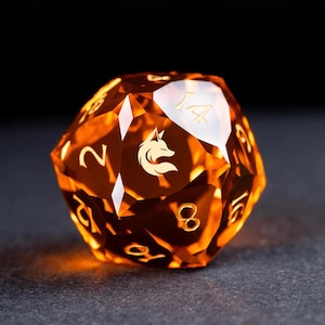 May include: A large, faceted, orange-colored D20 die with gold numbering and a gold fox logo. The die is made of a translucent material, with light shining through it. The numbers are clearly visible, and the fox logo is centered on one face.