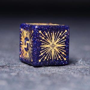 Op de afbeelding: Een blauwe en gouden dobbelsteen met een stervormig ontwerp. De dobbelsteen is gemaakt van een glinsterend materiaal.