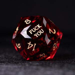 May include: A red, multi-sided die with gold numerals and the words "FUCK YOU" in gold lettering. The die is faceted and appears to be made of a translucent material, with a dark background.
