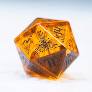 Op de afbeelding: Een twintigzijdige dobbelsteen in amber met zwarte markeringen. Elke zijde toont een nummer en een decoratief ontwerp. Deze dobbelsteen is ontworpen voor tabletop-rollenspellen.