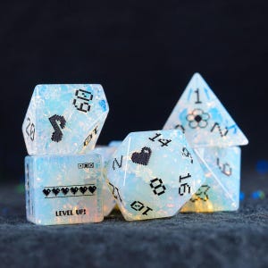 Könnte beinhalten: Sechs Stück iridescent blaue und weiße Würfel mit schwarzen pixelartigen Zahlen und Symbolen. Die Würfel sind in Form eines Würfels, einer Pyramide und eines Dodekaeders. Die Würfel haben den Text "LEVEL UP!" auf einer Seite.