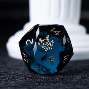 May include: A dark teal D20 die with gold numbering and a gold character illustration. The die is a gaming accessory. The die is set against a dark background with a white pillar in the background.