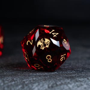 Puede incluir: Un dado rojo de veinte caras con números dorados y un emblema de dragón dorado. El dado se encuentra sobre un fondo oscuro y texturizado, resaltando su diseño y color.