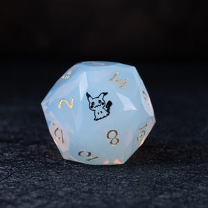 May include: A pale blue, multi-sided D20 die featuring gold numerals and a black Mimikyu design. The die is made from a translucent material. The numbers are in a serif font. Ideal for role-playing games.