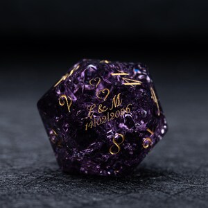 May include: A purple, multi-sided die with gold-coloured numbers and lettering. The die has the initials "L & M" and the date "14/02/2026" engraved on it, along with two heart symbols. The die is set against a dark background.