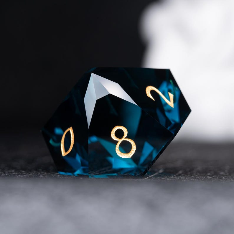 May include: A deep blue, faceted, gemstone dice with gold numbers 0, 2, and 8. The dice is resting on a dark surface.