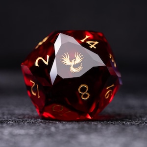 Op de afbeelding: Een facetgeslepen, rode D20 dobbelsteen met gouden cijfers en een gouden feniksembleem. De dobbelsteen is gemaakt van een doorschijnend materiaal, met scherpe randen en een gepolijste afwerking. De cijfers zijn duidelijk zichtbaar en het feniksembleem is gecentreerd op één zijde.