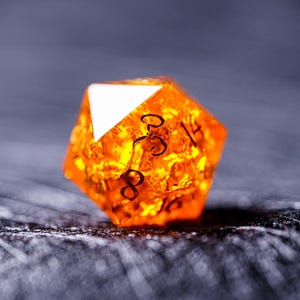 May include: A bright orange, translucent, 20-sided die with black numbers. The die is made of a clear material with a glittery finish.