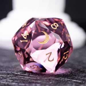 Op de afbeelding: Een roze, veelzijdige d20-dobbelsteen met gouden cijfers en een halve maan symbool. De dobbelsteen is gemaakt van een doorschijnend materiaal. De cijfers en het maansymbool zijn duidelijk zichtbaar. Ontworpen voor tabletop rollenspellen.