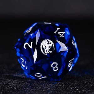 May include: A twenty-sided die, or D20, in a deep blue hue, featuring white numbering and a dragon emblem. The die is designed for role-playing games. It is made of a translucent material with a reflective surface.