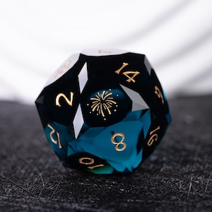 May include: A blue and black 12-sided die with gold numbers and a gold firework design. The die is made of glass and has a faceted design.